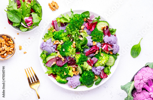 Yummy vegan salad with purple cauliflower  broccoli  fresh cucumbers  onion  lettuce with walnuts  white table background  top view