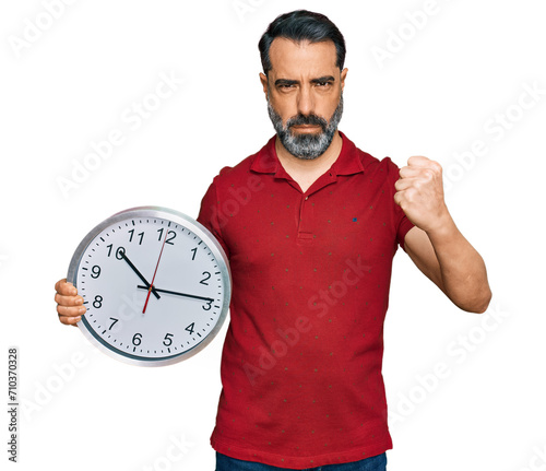 Middle aged man with beard holding big clock annoyed and frustrated shouting with anger, yelling crazy with anger and hand raised