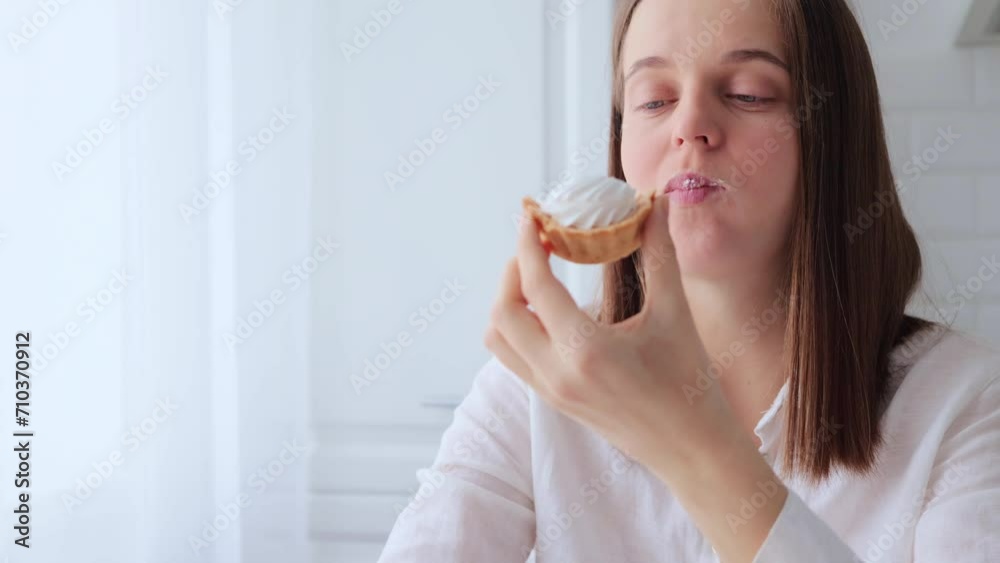 Satisfied brown haired woman wearing white shirt sitting in kitchen and eating delicious sugary cake while breaking diet having cheat meal enjoying delicious baking Satisfied brown haired woman wearing white shirt sitting in kitchen and eating delicious sugary cake while breaking diet having cheat meal enjoying delicious baking