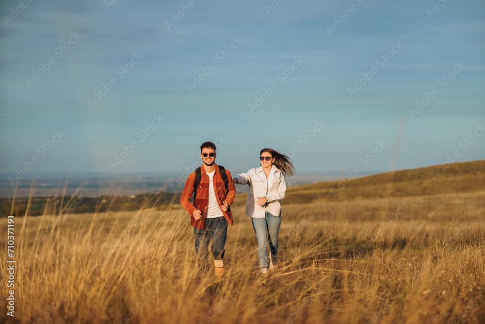 A couple is running on a big golden meadow, wearing sunglasses, smiling and enjoying the sunny autumn day.