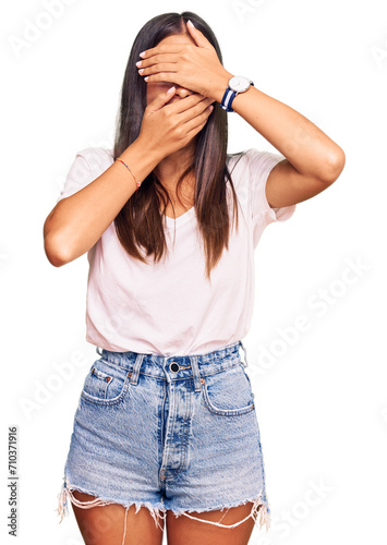 Young hispanic woman wearing casual white tshirt covering eyes and mouth with hands, surprised and shocked. hiding emotion