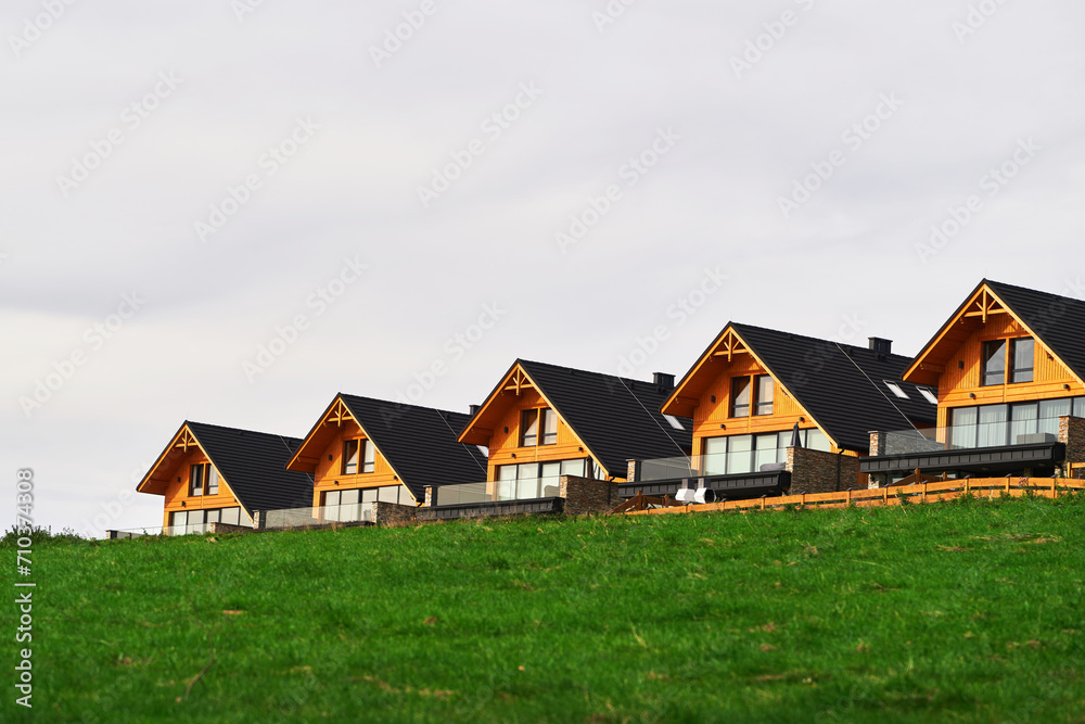 Modern wooden houses on a green hill with a stunning view of the sky and the fields. A row of modern wooden houses and green grass foreground. Modern wooden houses on a green hill with a stunning view of the sky and the fields. A row of modern wooden houses and green grass foreground.