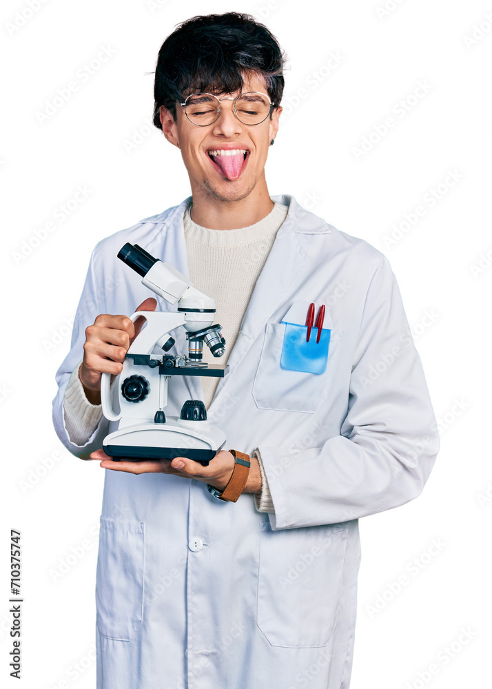 Handsome hipster young man wearing lab coat holding microscope sticking tongue out happy with funny expression. Handsome hipster young man wearing lab coat holding microscope sticking tongue out happy with funny expression.
