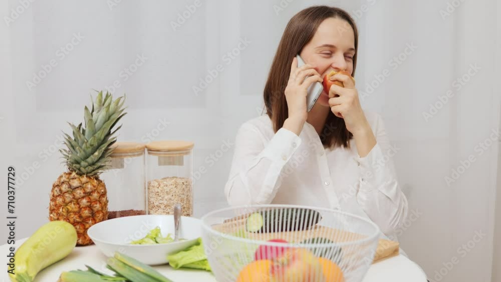 Beautiful woman eating apple and smartphone sitting in the kitchen at table and smiling while talking on mobile phone surrounded with organic food having pleasant conversation while cooking Beautiful woman eating apple and smartphone sitting in the kitchen at table and smiling while talking on mobile phone surrounded with organic food having pleasant conversation while cooking