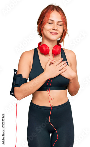 Young redhead woman wearing gym clothes and using headphones smiling with hands on chest with closed eyes and grateful gesture on face. health concept.
