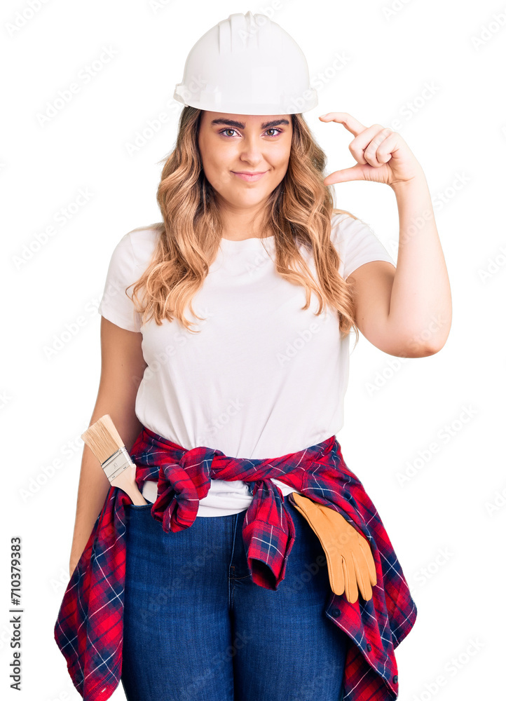 Young caucasian woman wearing security helmet smiling and confident gesturing with hand doing small size sign with fingers looking and the camera. measure concept. Young caucasian woman wearing security helmet smiling and confident gesturing with hand doing small size sign with fingers looking and the camera. measure concept.