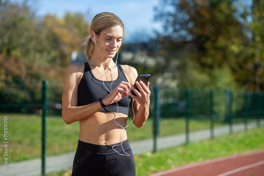 A fit female athlete in athletic wear selects music on her phone before a run in a sunny park setting. A fit female athlete in athletic wear selects music on her phone before a run in a sunny park setting.