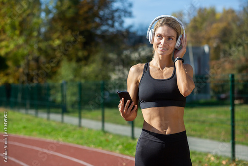 Athletic woman in workout gear using phone with headphones on sunny day at sports track. Fitness and technology concept.
