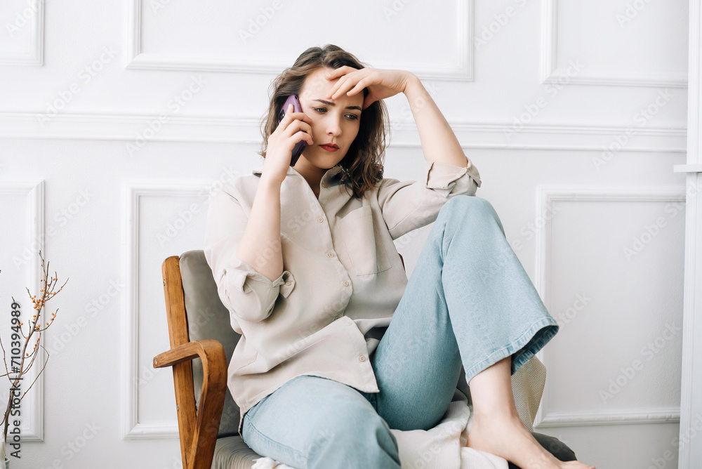 Unhappy Young Woman Receives Disheartening News During Phone Call, Speaks Emotionally While Seated Alone on an Armchair. Unhappy Young Woman Receives Disheartening News During Phone Call, Speaks Emotionally While Seated Alone on an Armchair.