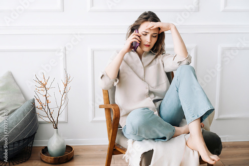 Upset Millennial Woman on Phone, Conveying Grief, Seated on Chair, Reacting to Unsettling News. Despaired sad millennial female calls by phone, talks, sits on chair, suffering from bad news.
