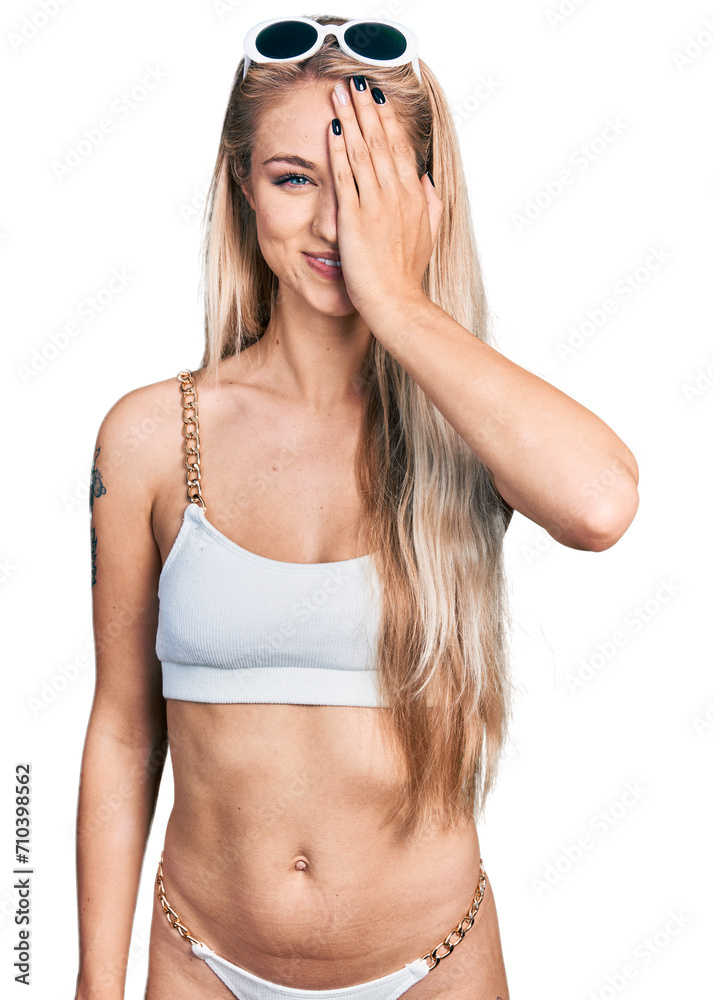Beautiful young blonde woman wearing bikini covering one eye with hand, confident smile on face and surprise emotion. Beautiful young blonde woman wearing bikini covering one eye with hand, confident smile on face and surprise emotion.