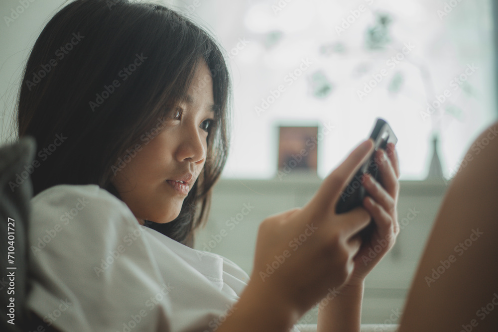 Happy Asian girl relaxing on a comfortable couch at home texting and messaging on a smartphone, smiling girl uses a cellphone, chatting online message, and shopping online from home. Happy Asian girl relaxing on a comfortable couch at home texting and messaging on a smartphone, smiling girl uses a cellphone, chatting online message, and shopping online from home.