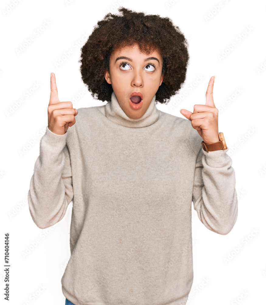 Young hispanic girl wearing wool winter sweater amazed and surprised looking up and pointing with fingers and raised arms. Young hispanic girl wearing wool winter sweater amazed and surprised looking up and pointing with fingers and raised arms.