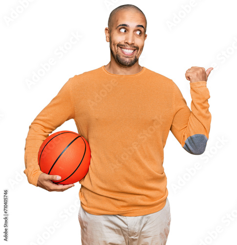Hispanic adult man holding basketball ball pointing thumb up to the side smiling happy with open mouth