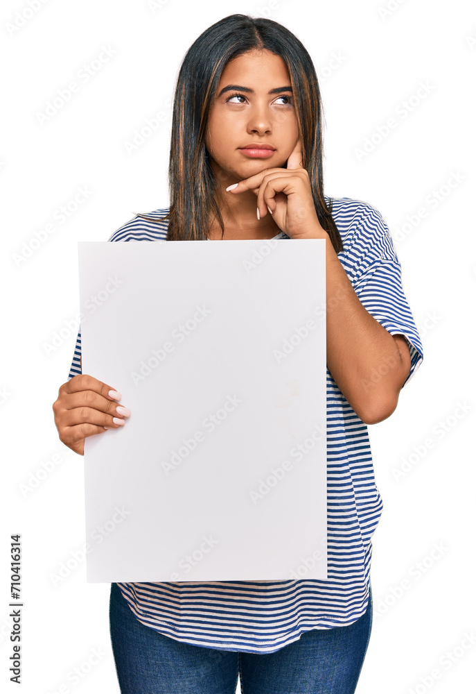 Young latin girl holding blank empty banner serious face thinking about question with hand on chin, thoughtful about confusing idea Young latin girl holding blank empty banner serious face thinking about question with hand on chin, thoughtful about confusing idea