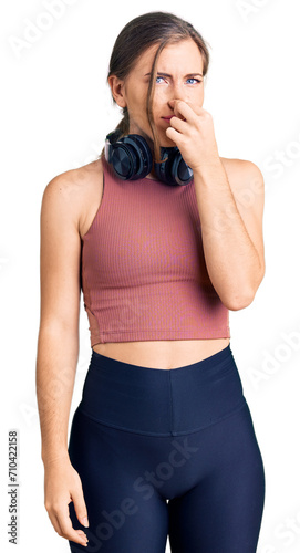 Beautiful caucasian young woman wearing gym clothes and using headphones smelling something stinky and disgusting, intolerable smell, holding breath with fingers on nose. bad smell