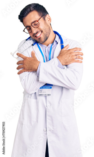 Young hispanic man wearing doctor uniform and stethoscope hugging oneself happy and positive, smiling confident. self love and self care