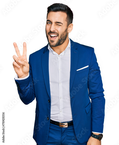 Young hispanic man wearing business jacket smiling with happy face winking at the camera doing victory sign with fingers. number two.