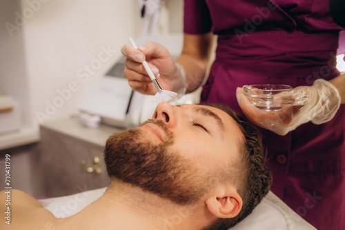 Beautician applying scrub onto young man's face in spa salon photo