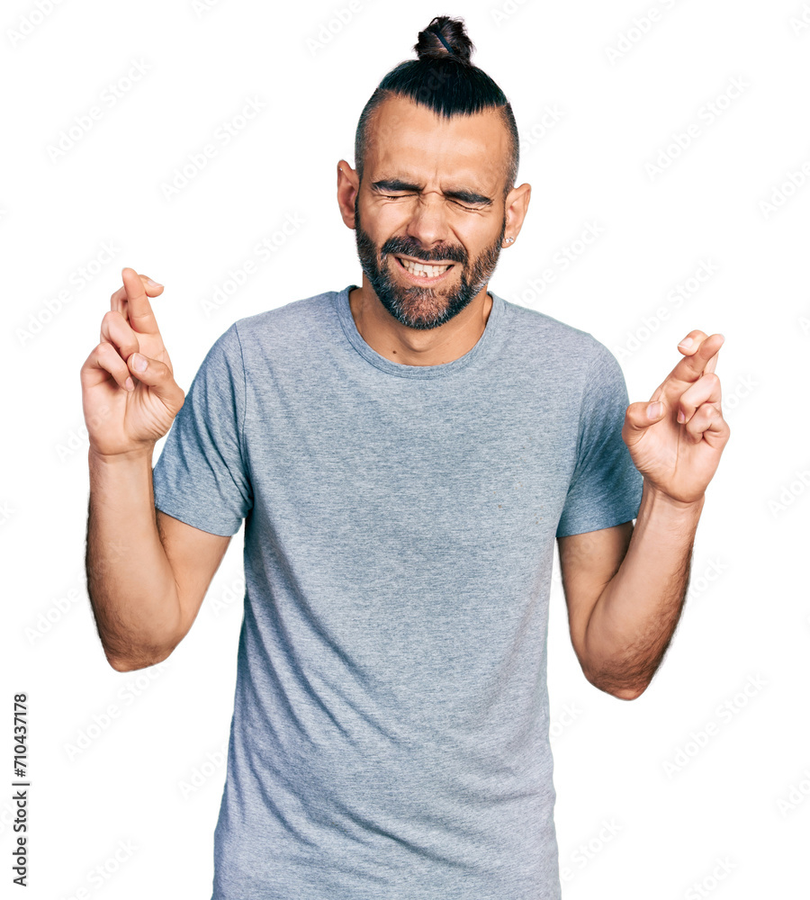 Hispanic man with ponytail wearing casual grey t shirt gesturing finger crossed smiling with hope and eyes closed. luck and superstitious concept. Hispanic man with ponytail wearing casual grey t shirt gesturing finger crossed smiling with hope and eyes closed. luck and superstitious concept.