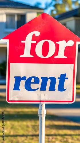 A vibrant red 'for rent' sign with bold white and blue lettering, prominently displayed on a stand with a softly blurred residential home in the background
