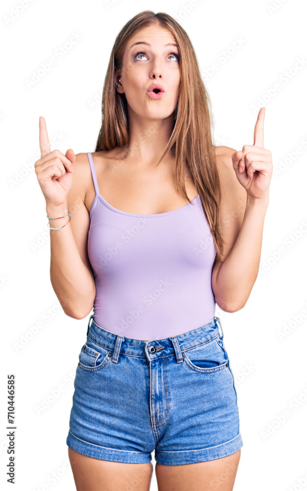 Young beautiful blonde woman wearing casual style with sleeveless shirt amazed and surprised looking up and pointing with fingers and raised arms. Young beautiful blonde woman wearing casual style with sleeveless shirt amazed and surprised looking up and pointing with fingers and raised arms.