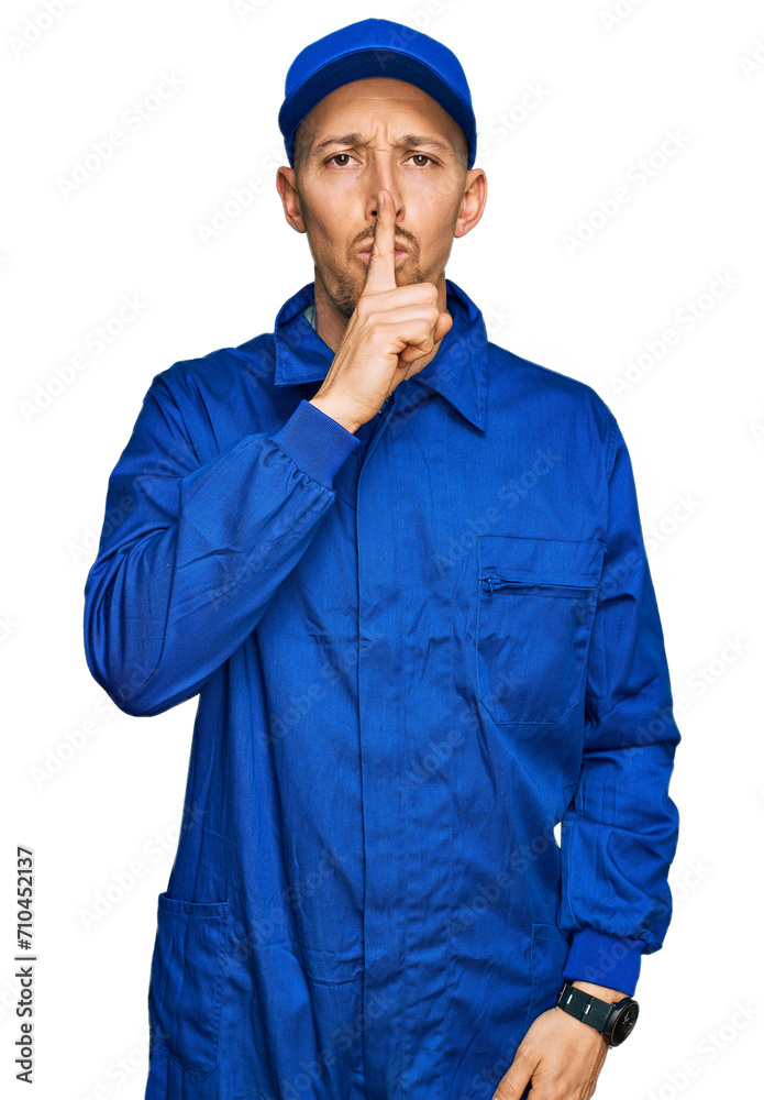 Bald man with beard wearing builder jumpsuit uniform asking to be quiet with finger on lips. silence and secret concept.