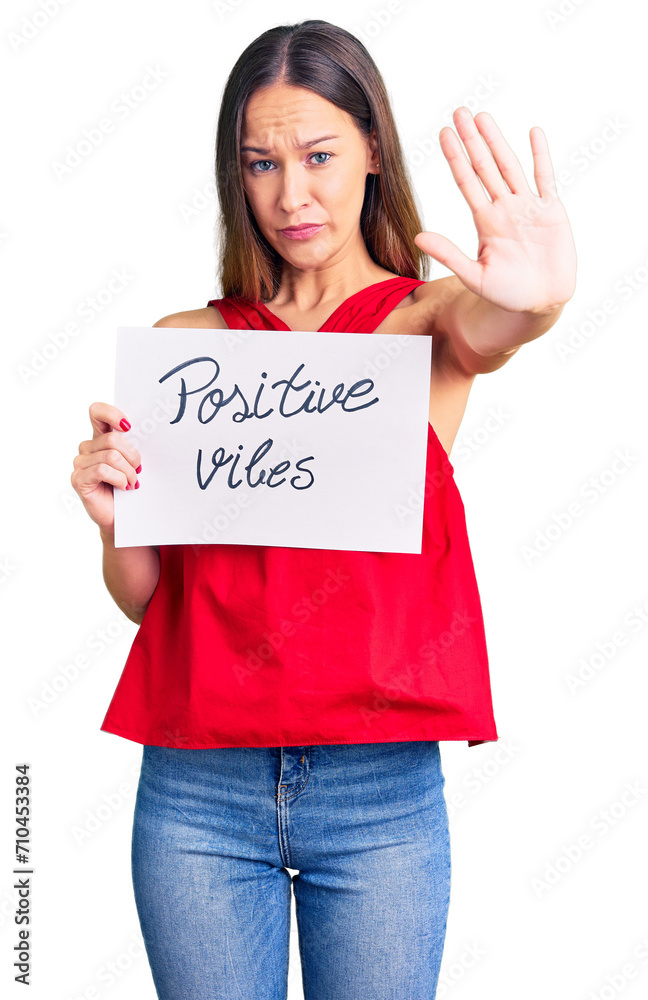 Beautiful brunette young woman holding positive vibes banner with open hand doing stop sign with serious and confident expression, defense gesture Beautiful brunette young woman holding positive vibes banner with open hand doing stop sign with serious and confident expression, defense gesture