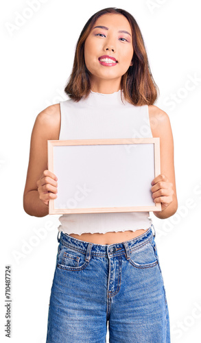 Young beautiful chinese girl holding empty white chalkboard looking positive and happy standing and smiling with a confident smile showing teeth