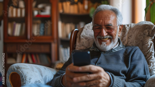 Close-up senior smiling relaxed retired man with beard and glasses sitting comfortably at home on armchair using mobile phone, communication concept photo