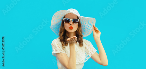 Portrait of beautiful young woman posing blowing her lips sending sweet air kiss wearing white summer straw hat on colorful blue background