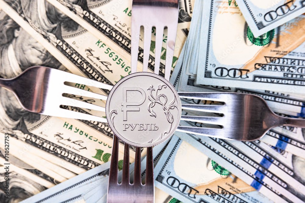 A new metal coin of the Russian ruble, with a denomination of one ruble against the background of American banknotes and metal forks. Crisis in Russia. - obrazy, fototapety, plakaty 
