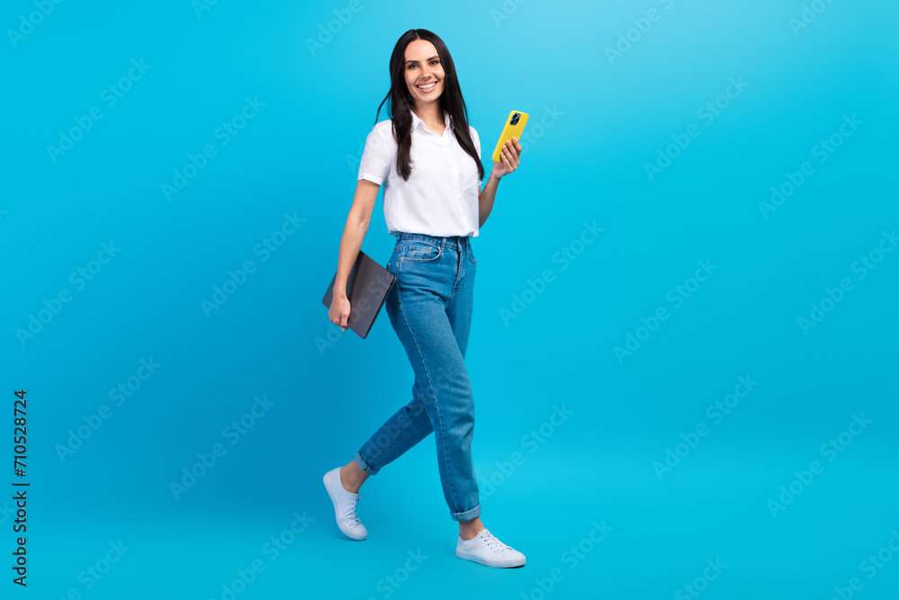 Full length photo of positive pretty girl dressed white shirt walking communicating modern gadget empty space isolated blue color background Full length photo of positive pretty girl dressed white shirt walking communicating modern gadget empty space isolated blue color background