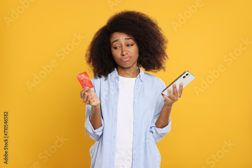 Confused woman with credit card and smartphone on orange background. Debt problem