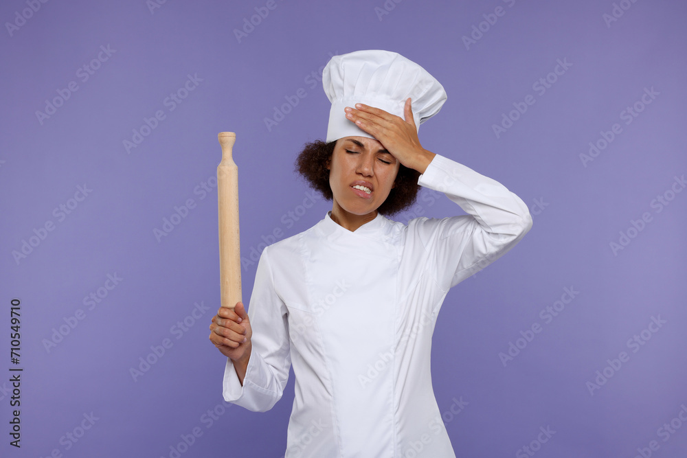 Frustrated female chef in uniform holding rolling pin on purple background Frustrated female chef in uniform holding rolling pin on purple background