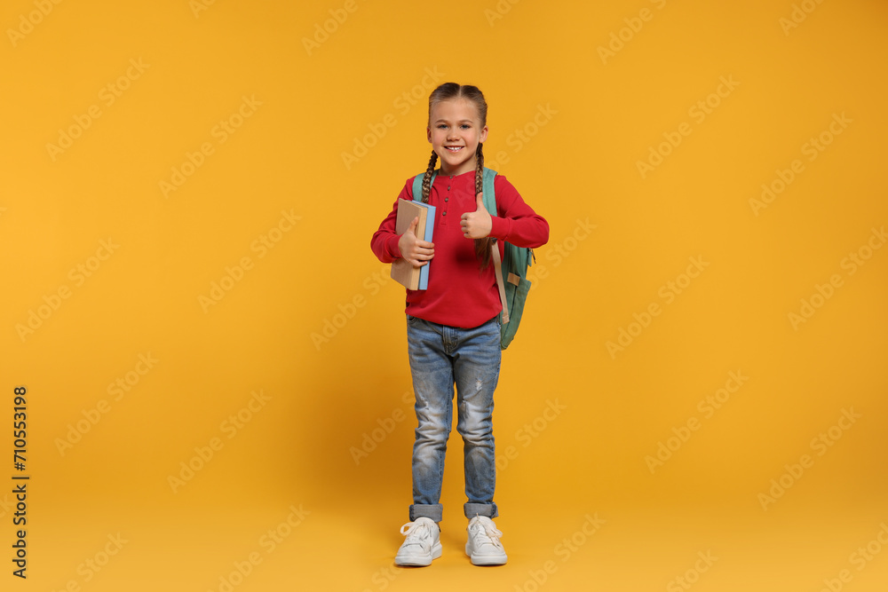 Happy schoolgirl with backpack and books showing thumb up gesture on orange background