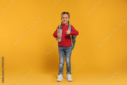 Happy schoolgirl with backpack and books showing thumb up gesture on orange background © New Africa