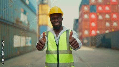 Happy African American industrial engineer in safety vest and hard hat standing and showing thumbs up to camera. Confident black foreman showing thumb up in approval, recommending, like smth good photo