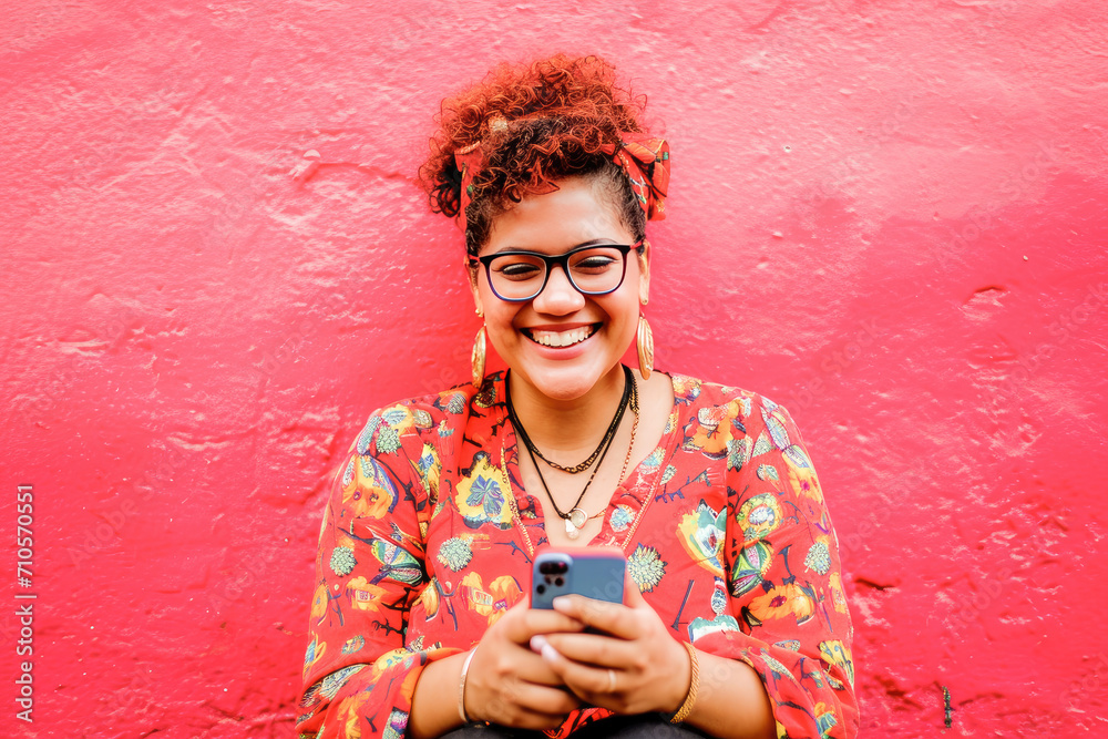 Fototapeta premium A vibrant portrait of a joyful young woman smiling while texting on her mobile phone against a vivid red background.