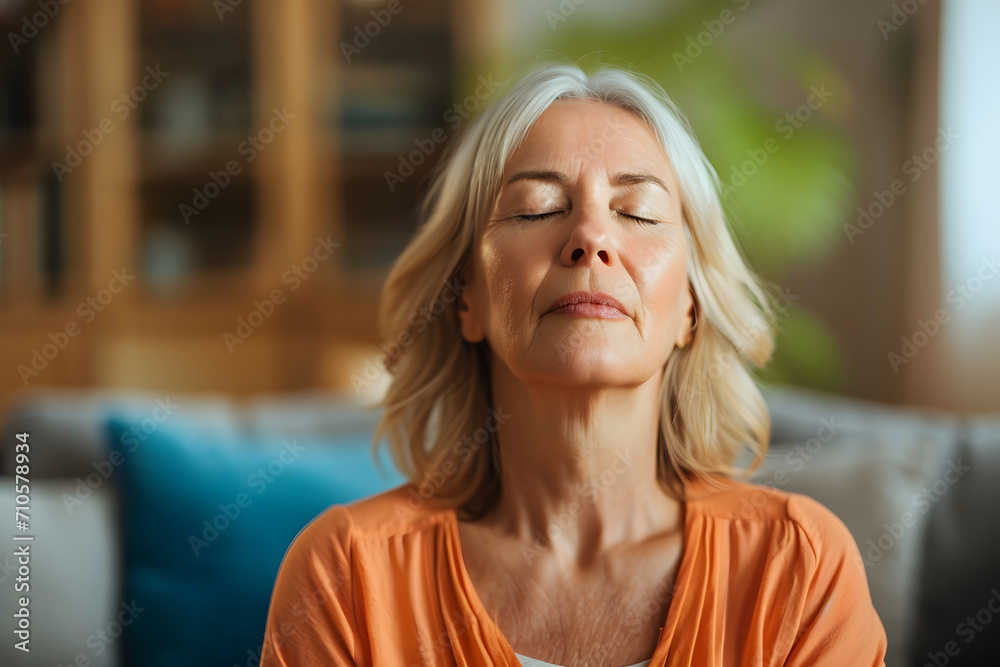 At home, a middle-aged woman closes her eyes in contemplation and relaxes her body and mind in her living room. Mental health and meditation for a stress-free concept. Self-care and well-being.