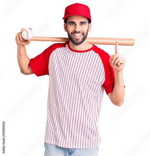 Young handsome man with beard playing baseball holding bat and ball surprised with an idea or question pointing finger with happy face, number one