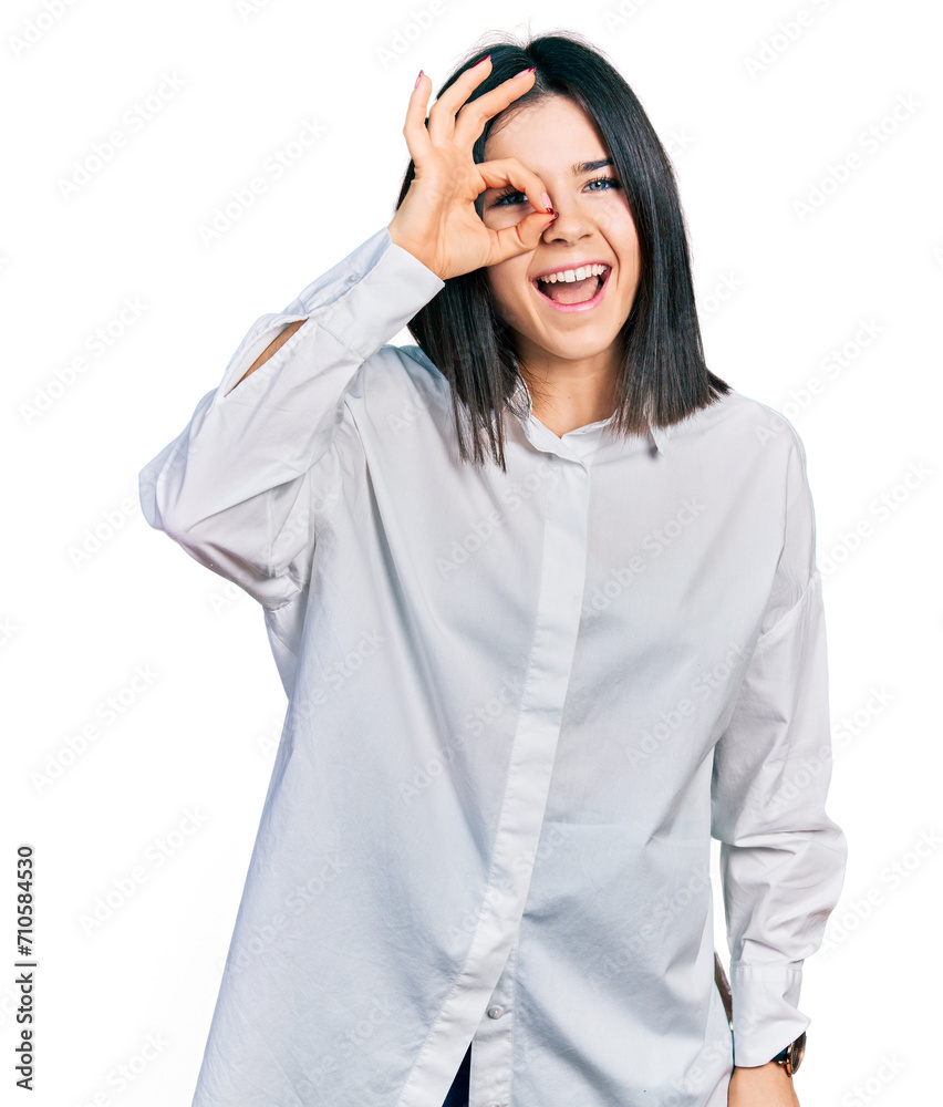 Young brunette woman with blue eyes wearing oversize white shirt doing ok gesture with hand smiling, eye looking through fingers with happy face. Young brunette woman with blue eyes wearing oversize white shirt doing ok gesture with hand smiling, eye looking through fingers with happy face.