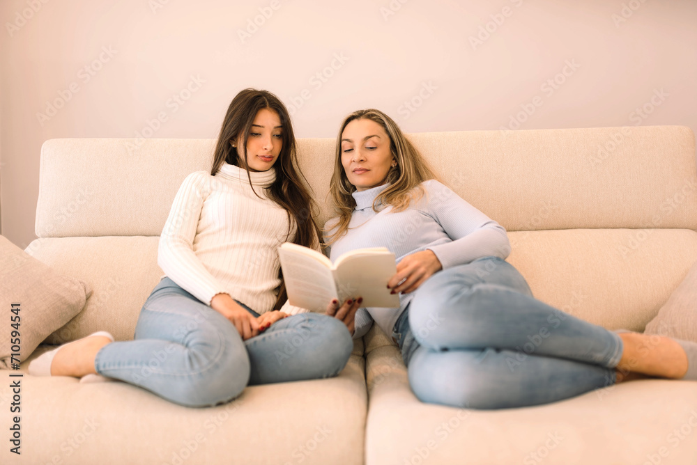 mother and teenage daughter sitting on couch at home reading a book together