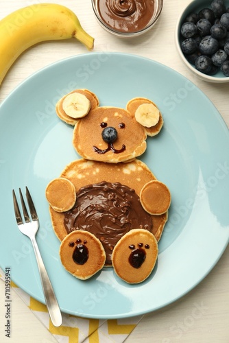 Creative serving for kids. Plate with cute bear made of pancakes, blueberries, bananas and chocolate paste on light wooden table, flat lay