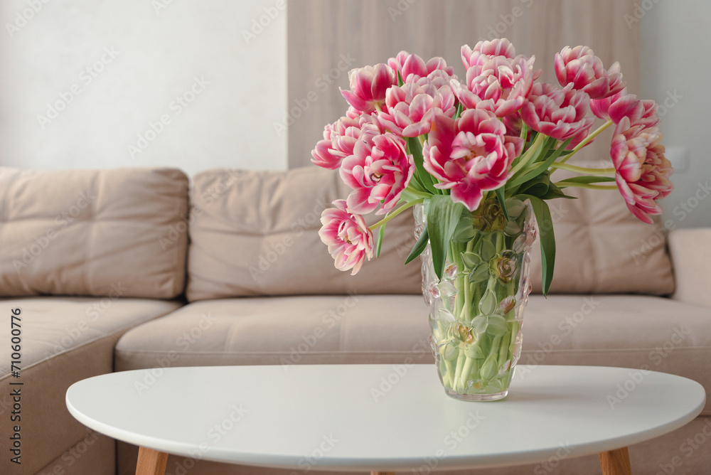Vase of pink tulips on coffee table with blurred background of beige modern cozy light living room.  Copy space