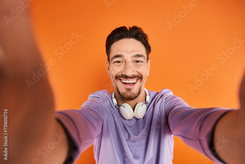 happy bearded man in wireless headphones listening music and looking at camera on orange background