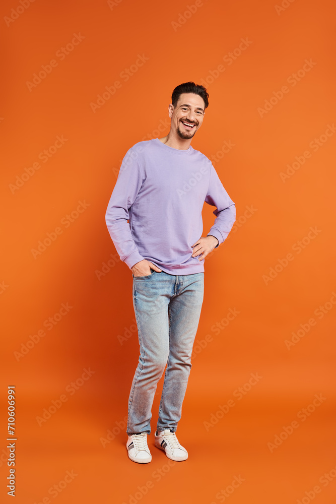 happy man in purple sweater and jeans standing with hand in pocket on orange background, casual wear happy man in purple sweater and jeans standing with hand in pocket on orange background, casual wear