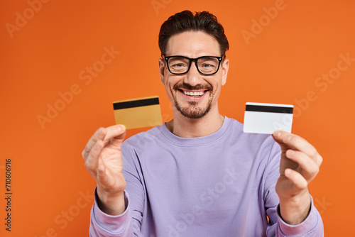 cheerful bearded man in glasses holding credit cards on orange background, shopping and consumerism