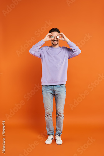 joyful man holding bitcoins over his eyes and smiling at camera on orange background, cryptocurrency