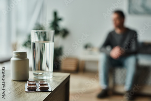 medication in bottle and blister pack near glass of water on table and blurred man on background